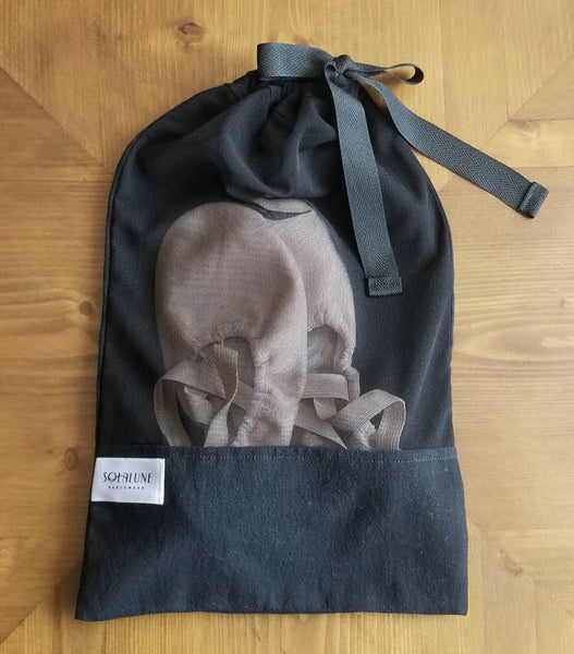 Black drawstring bag on a wooden table. The top three-quarters of the front of the bag is sheer black mesh, and the bottom quarter is a solid black cotton panel. A white satin brand logo label reading "Solalune Dancewear" is sewn at the top left of the cotton panel. The opening at the top of the bag is drawn closed into a bow with a wide, flat black tie. Pink ballet slippers are visible inside the bag through the black mesh panel.