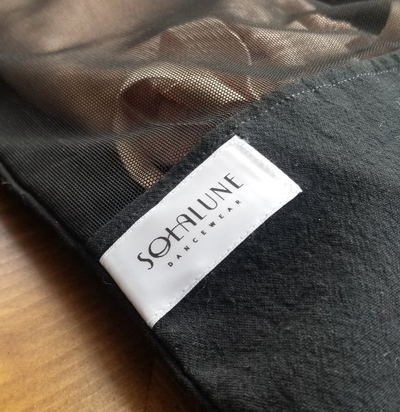 Detail view of the white satin brand logo label reading "Solalune Dancewear" stitched onto the black cotton panel of the drawstring bag. Pink ballet slippers are visible above through the black sheer mesh.