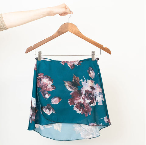 An opaque teal classic ballet wrap skirt with a painterly floral print in shades of soft pinks, burgundy, and white on a wooden clip hanger.