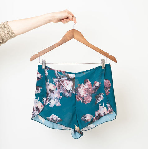 Pair of opaque teal shorts with a painterly floral print in shades of soft pinks, burgundy, and white on a wooden clip hanger.