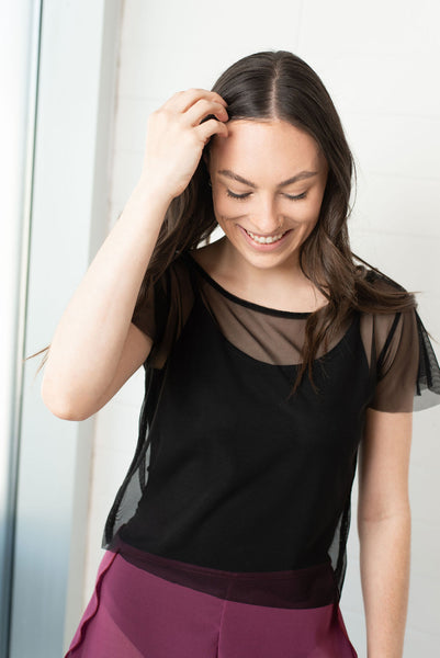 Dancer standing and smiling with eyes cast downward, wearing a black sheer mesh short sleeved t-shirt layered over a black leotard, with a pair of sheer raspberry coloured chiffon shorts. 