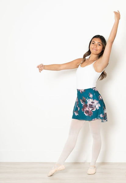 Smiling dancer in tendu with arms in fourth position, wearing nn opaque teal classic ballet wrap skirt with a painterly floral print in shades of soft pinks, burgundy, and white layered over a white leotard, pink tights and ballet slippers.